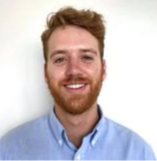 Head-and-shoulders portrait of a smiling man with red hair and a beard, wearing a light blue button-down shirt, standing against a white background.