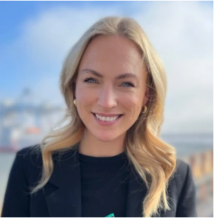 Head-and-shoulders portrait of a smiling blonde woman in a black blazer outdoors by the water.