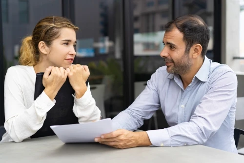 Two colleagues sit at a table outside an office, reviewing documents and discussing a project.