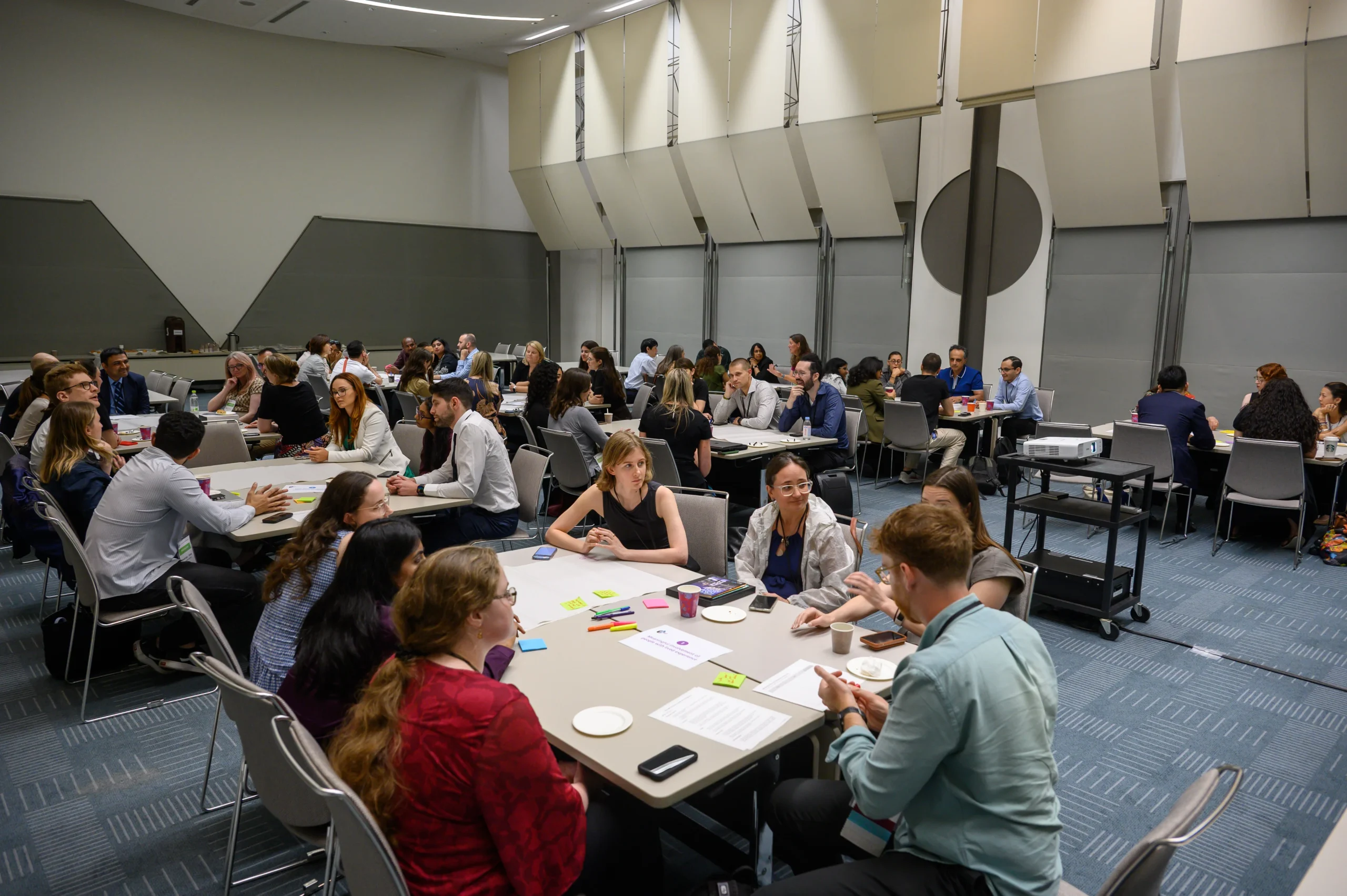 Group of professionals seated at round tables in a large conference room, engaged in discussion and notes during a workshop.