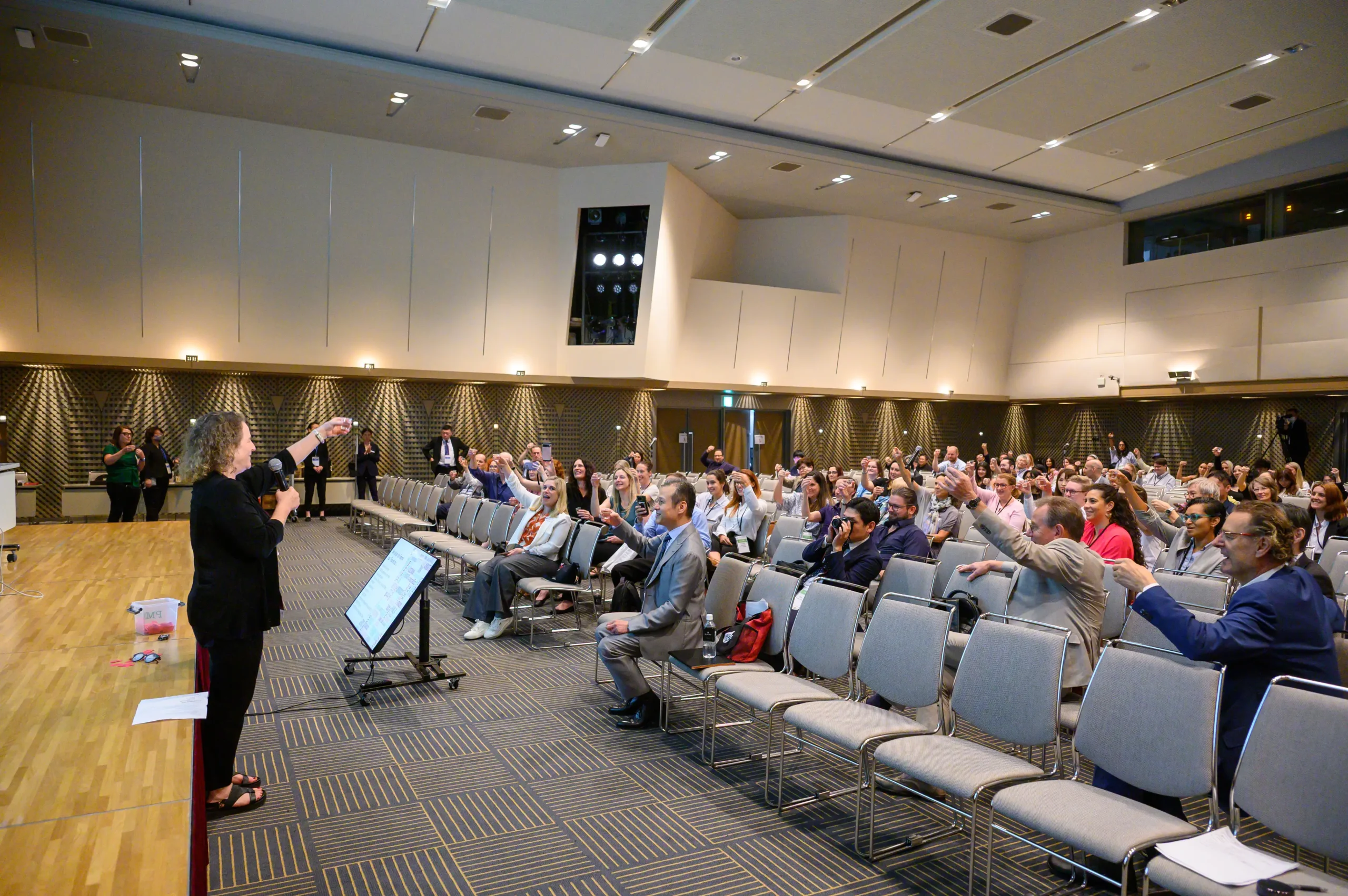 Speaker at a podium addressing a packed conference hall as audience members raise their glasses in a toast.