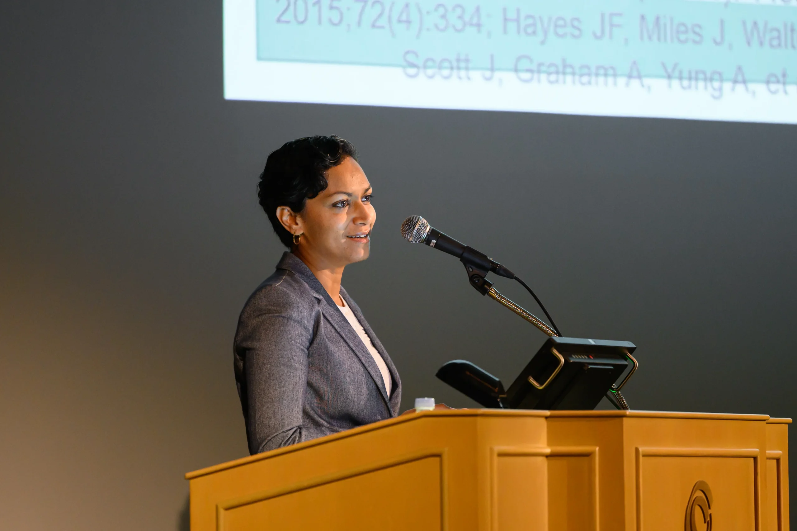 Woman speaking at a wooden podium with a microphone in front of a projected slide behind her, in a conference setting.