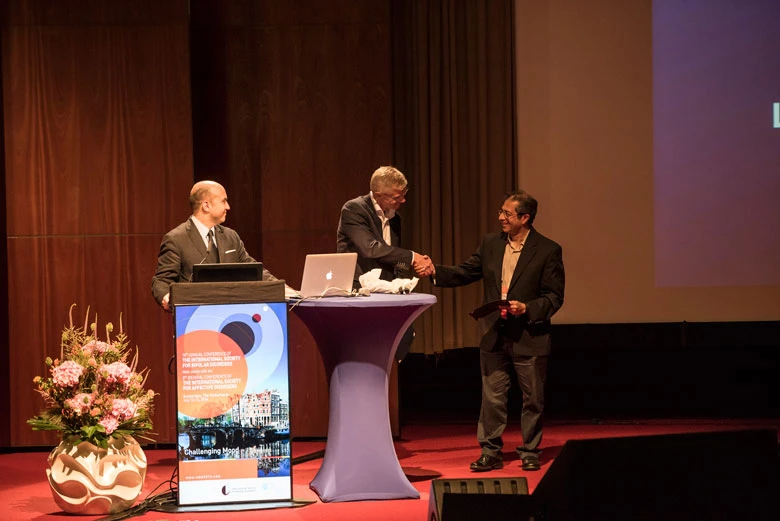 Three men on a conference stage; two shake hands at a purple-topped table while a third man stands at a podium with a poster and a laptop nearby.