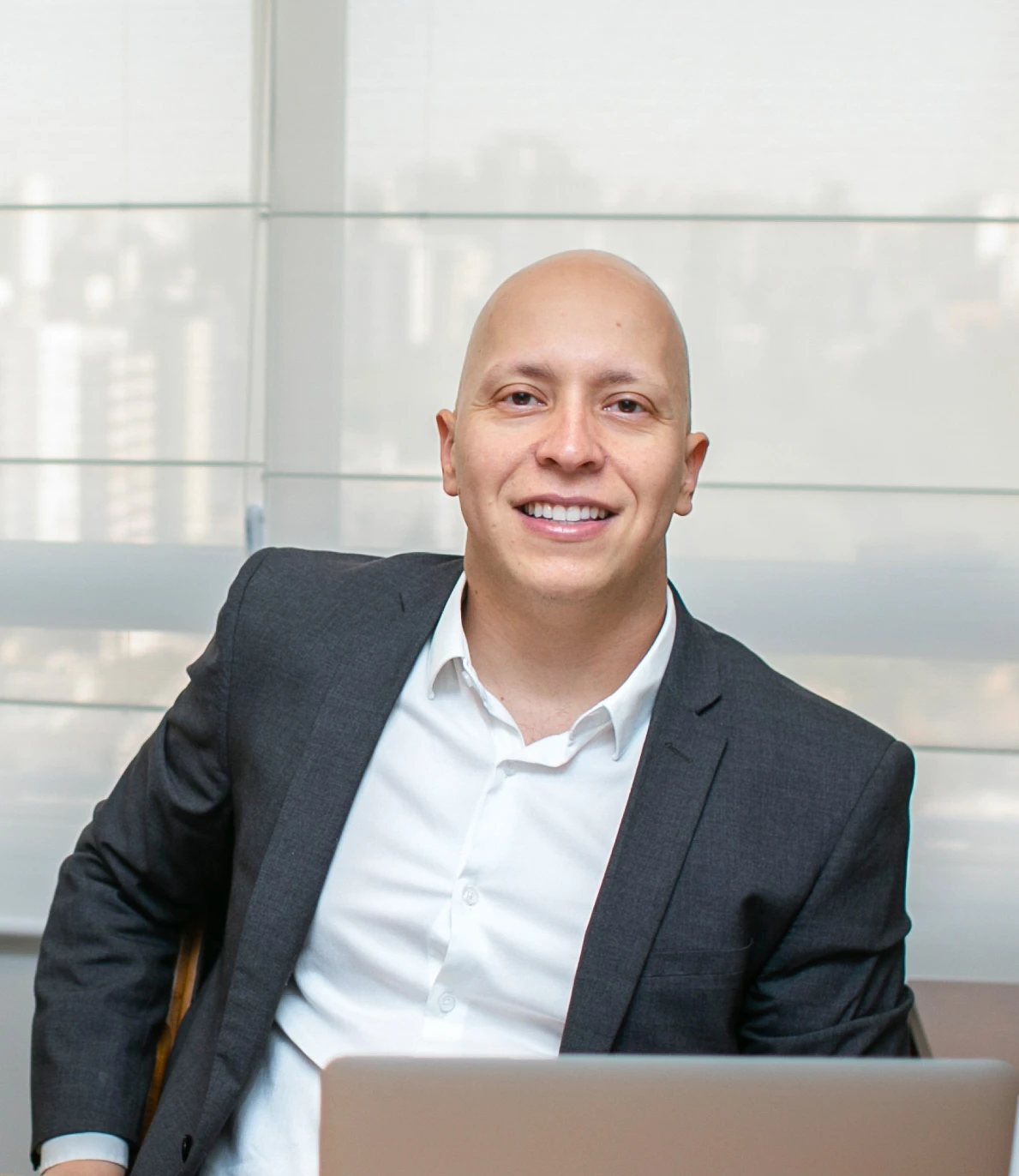 Smiling bald man in a dark suit and white shirt seated at a desk with a laptop in an office setting.