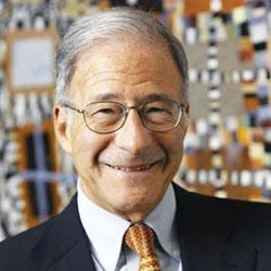 Smiling older man with glasses in a dark suit and orange tie, indoor background.