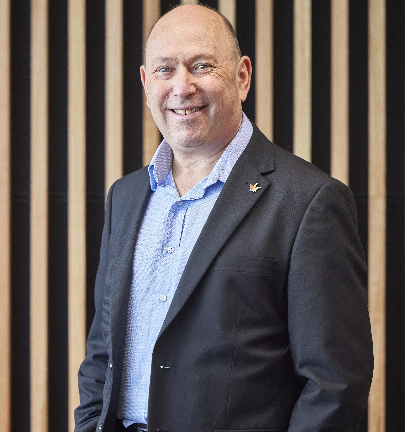 Smiling man in a blue shirt and dark blazer, standing in front of a wooden slat background.