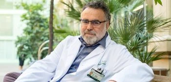 Senior doctor wearing a white coat and ID badge sits in a plant-filled lounge area, looking at the camera.