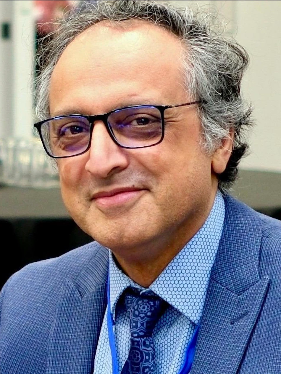 Smiling man with gray hair and glasses in a blue suit and patterned tie, indoors.