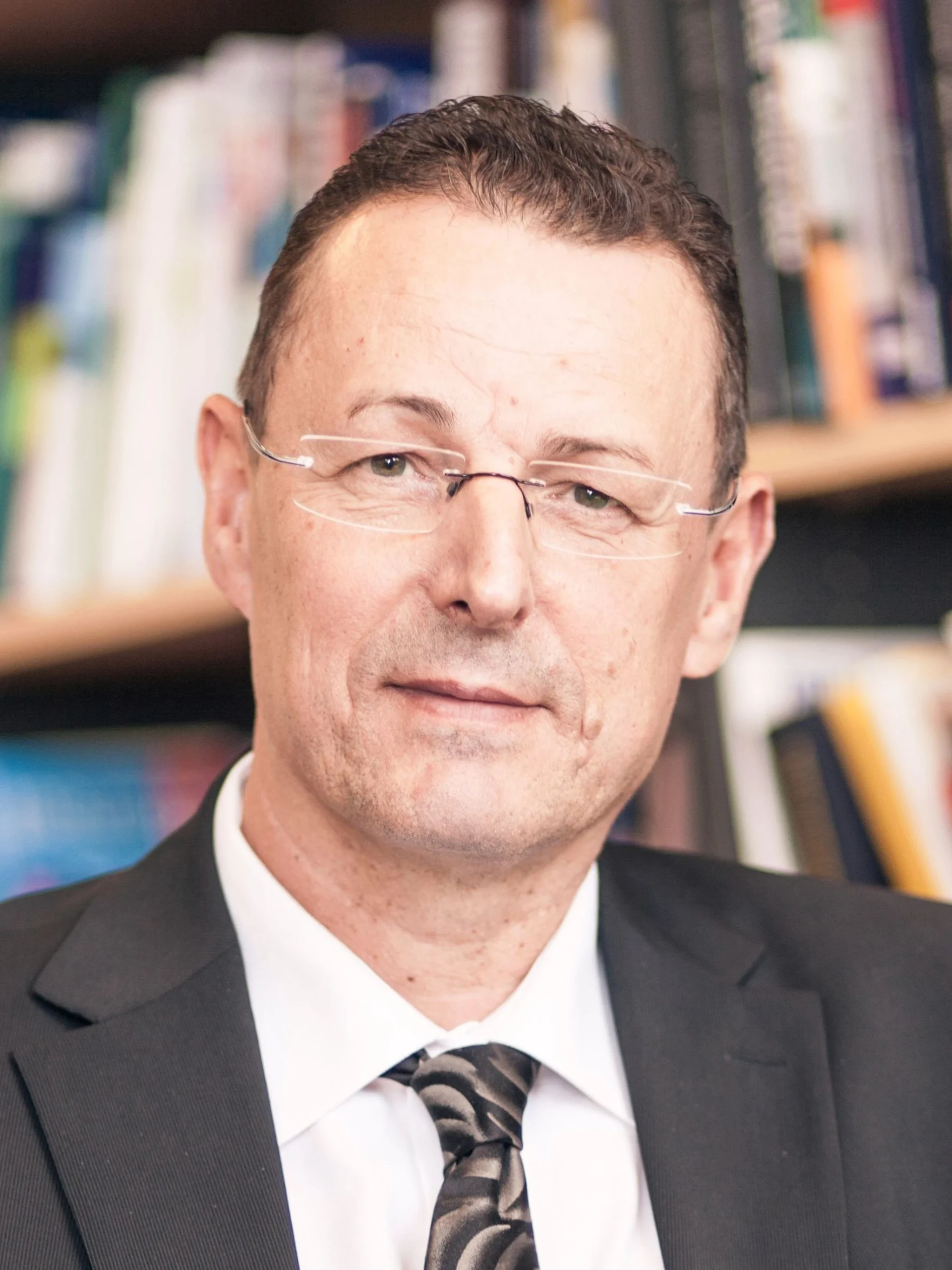 Portrait of a man in a suit and tie wearing thin glasses, in a library backdrop of bookshelves behind him.]","Close-up of a middle-aged man with short dark hair and wire-frame glasses, smiling slightly, with bookshelves in the background.","Professional headshot of a man in a dark suit and patterned tie, in a library setting.","Businessman wearing glasses and a suit, posed in front of bookshelves in an indoor library."