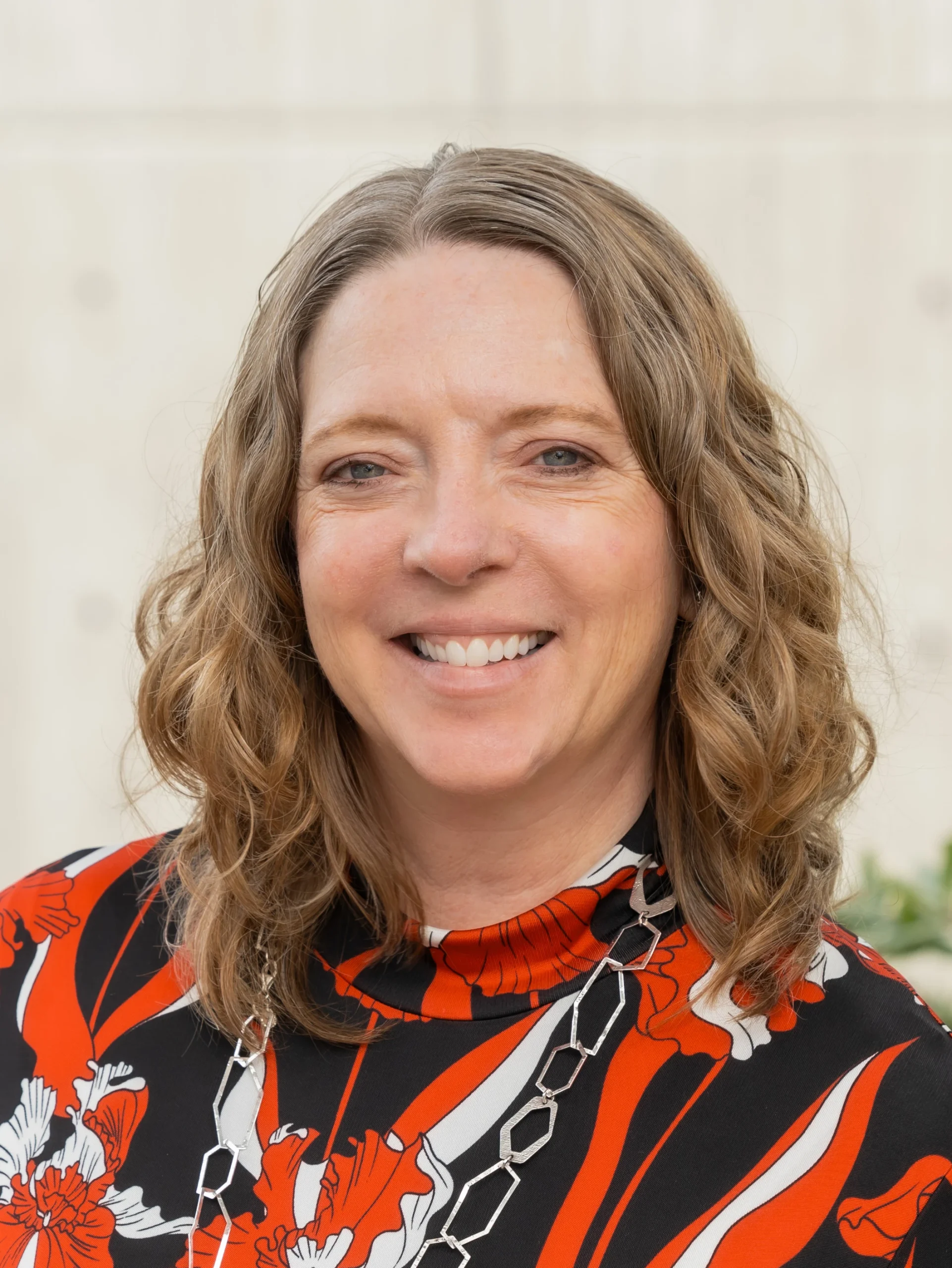Close-up of a smiling woman with wavy light brown hair wearing a red and black floral dress and a silver necklace.