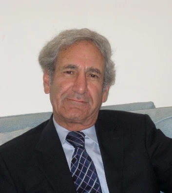 Portrait of an older man in a dark suit and striped tie, seated on a light blue sofa against a pale wall.