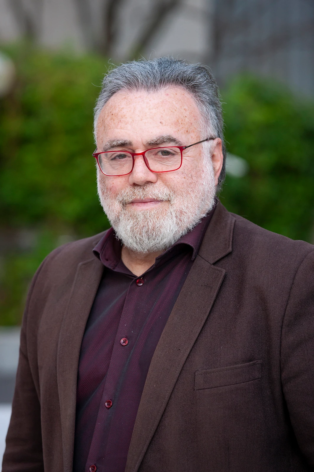 Portrait of an older man with gray hair and beard, red glasses, wearing a burgundy shirt and brown blazer outdoors, looking at the camera.
