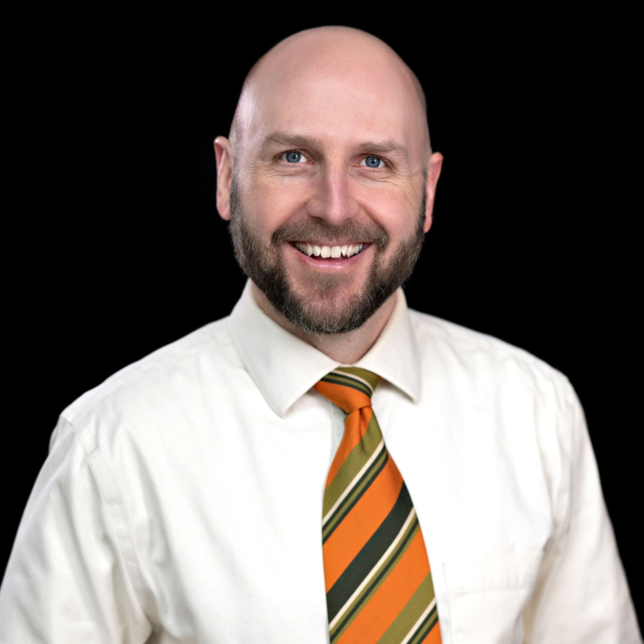 Close-up of a smiling man in a white shirt and striped orange-green tie against a black background, cropped at the shoulders.]','